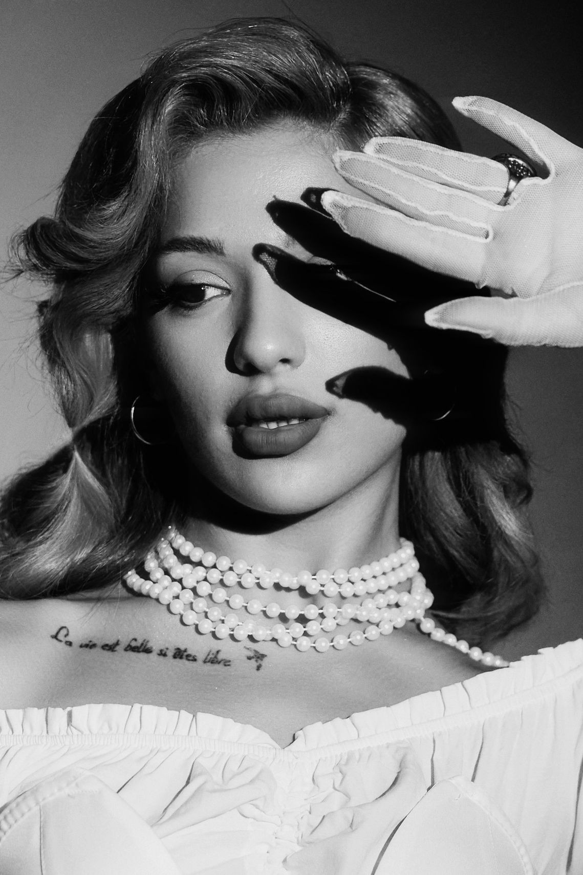 Black and white portrait of a woman with pearls, wearing gloves, shielding her eyes from light.